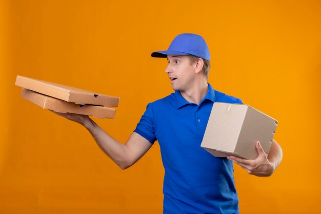 Young handsome delivery man in blue uniform and cap holding box package and pizza boxes looking aside surprised standing over orange wall