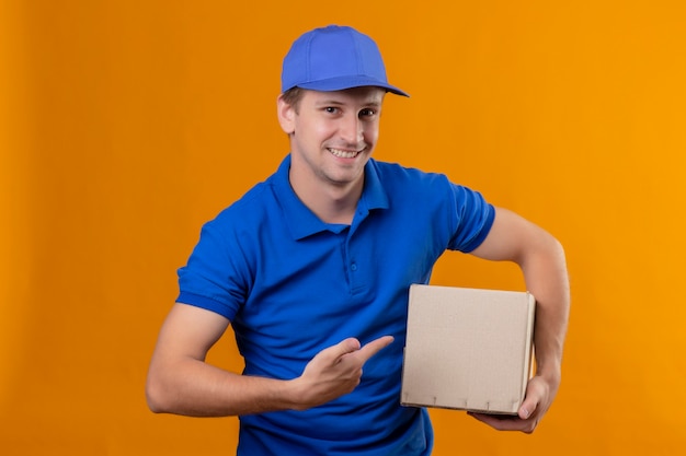 Молодой красавец-доставщик в синей форме и кепке держит коробку, указывая пальцем на нее