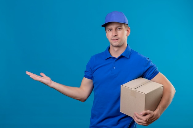 青い制服を着た若いハンサムな配達人と青い壁の上に立って笑みを浮かべて彼の手のコピースペースの腕を提示するボックスパッケージを保持しているキャップ