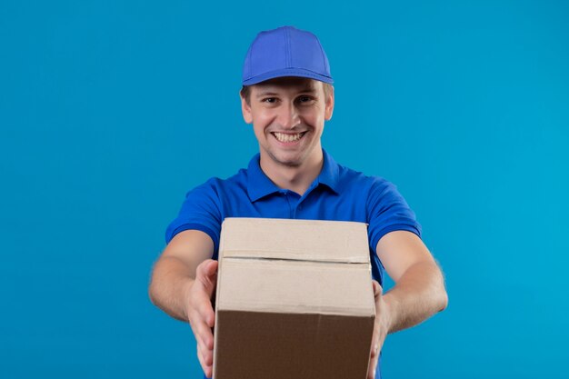 Young handsome delivery man in blue uniform and cap holding box package smiling cheerfully