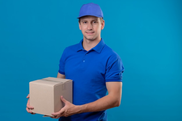 Free photo young handsome delivery man in blue uniform and cap holding box package smiling friendly happy and positive standing over blue wall