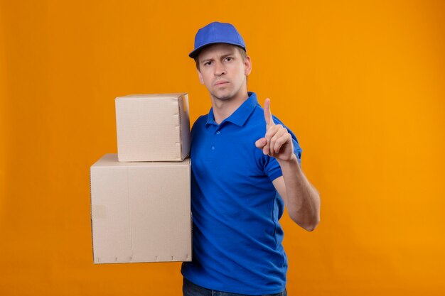 Young handsome delivery man in blue uniform and cap holding cardboard boxes pointing finger up warning with serious expression on face standing over orange wall