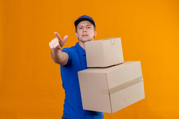 Молодой красивый курьер в синей форме и кепке держит картонные коробки, указывая на что-то пальцем