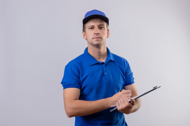 Young handsome delivery man in blue uniform and cap holding clipboard with pen looking confident standing over white wall