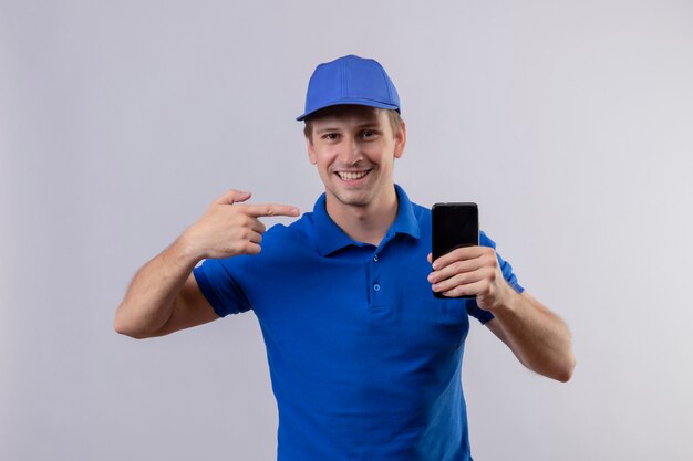 Молодой красивый курьер в синей форме и кепке держит мобильный телефон, указывая пальцем на него, дружелюбно улыбаясь, стоя над белой стеной