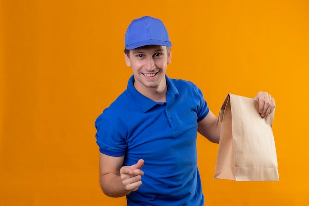 Young handsome delivery man in blue uniform and cap holding paper package pointing with finger to camera smiling friendly positive and happy standing over orange wall