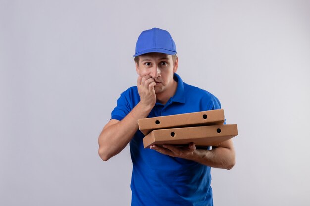 青い制服を着た若いハンサムな配達人とピザの箱を保持しているキャップが強調され、緊張して白い壁の上に立っている彼の爪をかむ