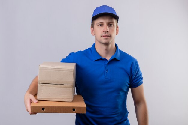 Молодой красивый курьер в синей форме и кепке, держащий коробки для пиццы с уверенным выражением лица, стоящий над белой стеной