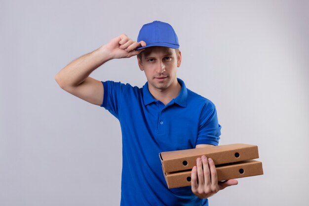 Young handsome delivery man in blue uniform and cap holding pizza boxes with confident expression on face standing over white wall