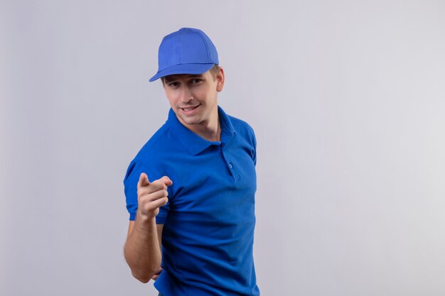 Young handsome delivery man in blue uniform and cap pointing to camera with index finger looking confident standing over white wall