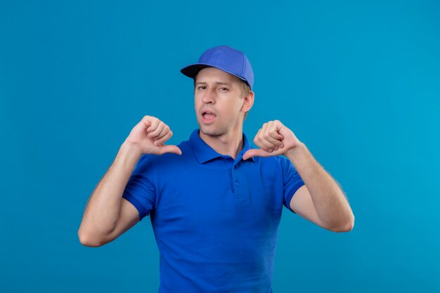 Free photo young handsome delivery man in blue uniform and cap pointing to himself with both hands with thumbs looking confident self-satisfied and proud standing over blue wall