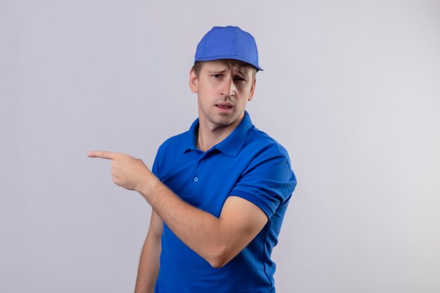 青い制服を着た若いハンサムな配達人と人差し指で指している側に白い壁を混乱して立っている探しているキャップ