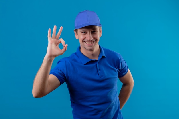 Giovane uomo di consegna bello in uniforme blu e cappuccio che sorride allegramente facendo segno giusto in piedi sopra la parete blu