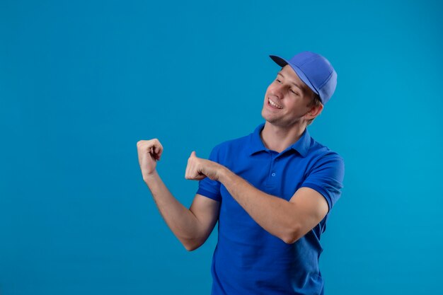 Молодой красивый курьер в синей униформе и кепке, весело улыбаясь, указывая в ответ с большими пальцами руки, стоя над синей стеной