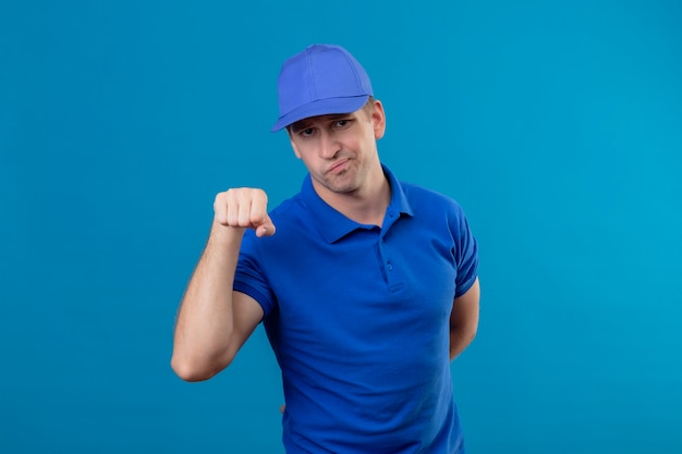 Молодой красивый курьер в синей форме и кепке со скептической улыбкой, сжимающий кулак в знак приветствия, стоящий над синей стеной