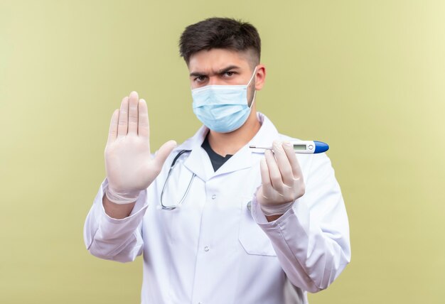 Молодой красивый доктор в синей медицинской маске, белом медицинском халате, белые медицинские перчатки и стетоскоп, сердито держит в руках электронный термометр, показывающий знак остановки