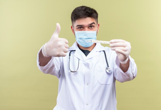 Молодой красивый врач в синей медицинской маске, белом медицинском халате, белые медицинские перчатки и стетоскоп, держащий термометр, делает счастливые пальцы вверх, стоя над стеной цвета хаки
