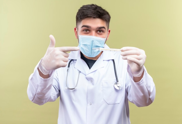 Young handsome doctor wearing blue medical mask white medical gown white medical gloves and stethoscope pointing to thermometer standing over khaki wall