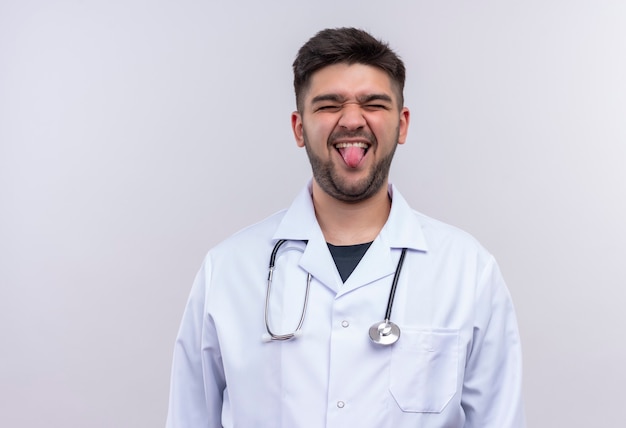 흰 벽 위에 서있는 그의 혀를 장난스럽게 보여주는 흰색 의료 가운과 청진기를 착용하는 젊은 잘 생긴 의사