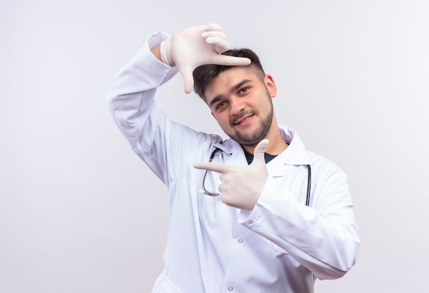 Young handsome doctor wearing white medical gown white medical gloves and stethoscope catching the frame standing over white wall