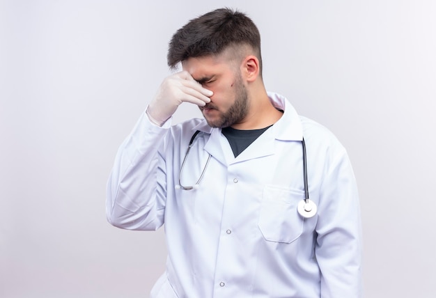 Молодой красивый врач в белом медицинском халате, белые медицинские перчатки и стетоскоп, закрывающий его насморк, стоя над белой стеной