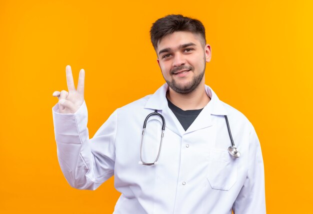 白い医療用ガウン白い医療用手袋と聴診器を身に着けている若いハンサムな医師は、オレンジ色の壁の上に立って喜んで見ている指でピースサインをしています