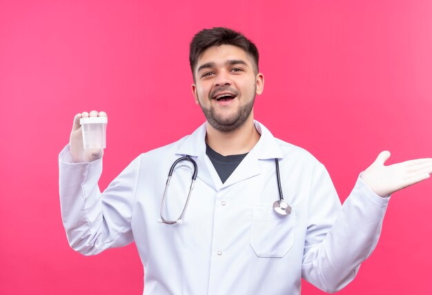 흰색 의료 가운 흰색 의료 장갑과 청진기를 입고 젊은 잘 생긴 의사는 분홍색 벽 위에 서있는 투명한 분석 컨테이너의 결과를 기뻐합니다.