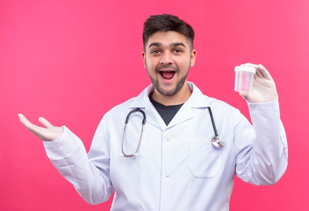 Young handsome doctor wearing white medical gown white medical gloves and stethoscope glad for results in transparent analysis container standing over pink wall