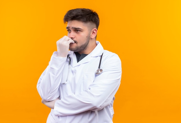 Young handsome doctor wearing white medical gown white medical gloves and stethoscope looking besides scared standing over orange wall