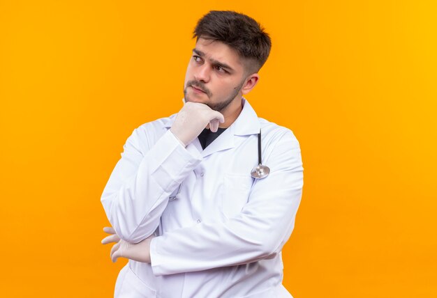 Young handsome doctor wearing white medical gown white medical gloves and stethoscope looking besides thoughtfully standing over orange wall