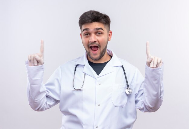 Young handsome doctor wearing white medical gown white medical gloves and stethoscope looking pointing up with forefingers standing over white wall