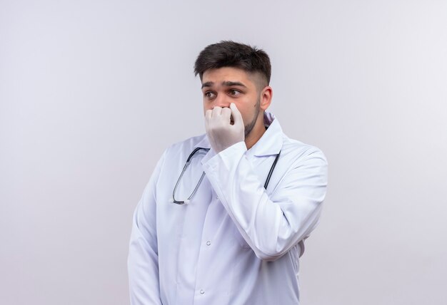 Young handsome doctor wearing white medical gown white medical gloves and stethoscope looking scared besides holding fist on mouse standing over white wall