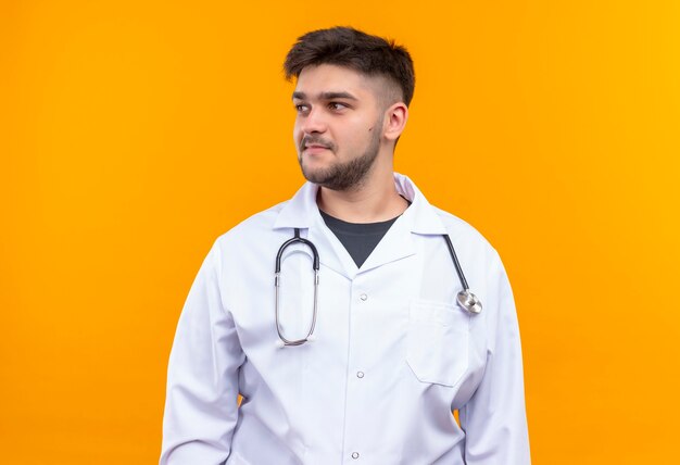 Young handsome doctor wearing white medical gown white medical gloves and stethoscope looking smily besides standing over orange wall