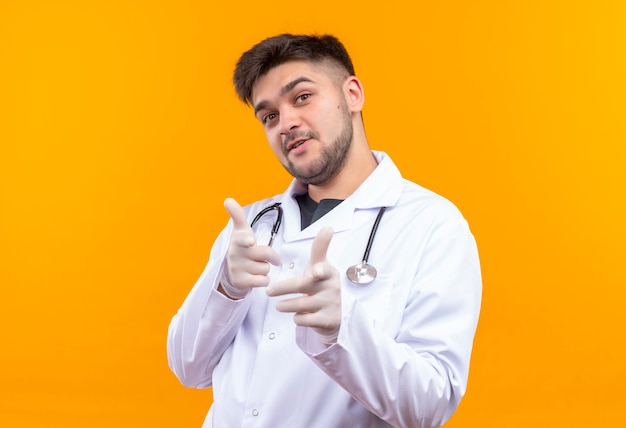 Young handsome doctor wearing white medical gown white medical gloves and stethoscope pointing happily on camera standing over orange wall