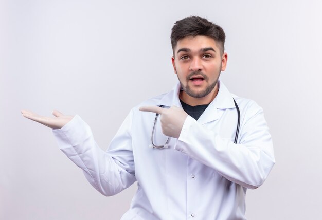 흰색 의료 가운 흰색 의료 장갑과 흰 벽 위에 서있는 오른손에 가리키는 청진기를 입고 젊은 잘 생긴 의사