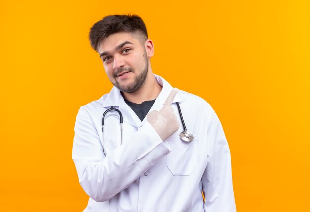 흰색 의료 가운 흰색 의료 장갑과 오렌지 벽 위에 서있는 집게 손가락으로 가리키는 청진기를 입고 젊은 잘 생긴 의사