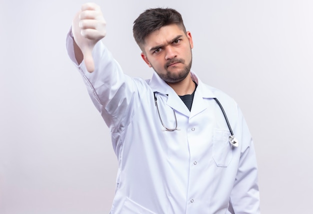白い医療用ガウン白い医療用手袋と聴診器を身に着けている若いハンサムな医者は、白い壁の上に立って悪い親指を非難している