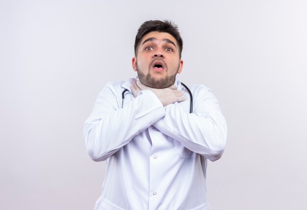 Young handsome doctor wearing white medical gown white medical gloves and stethoscope scared of death choking himself standing over white wall