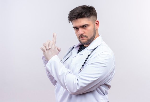 Молодой красивый врач в белом медицинском халате, белые медицинские перчатки и стетоскоп, серьезно глядя, делает пистолет с руками, стоящими над белой стеной
