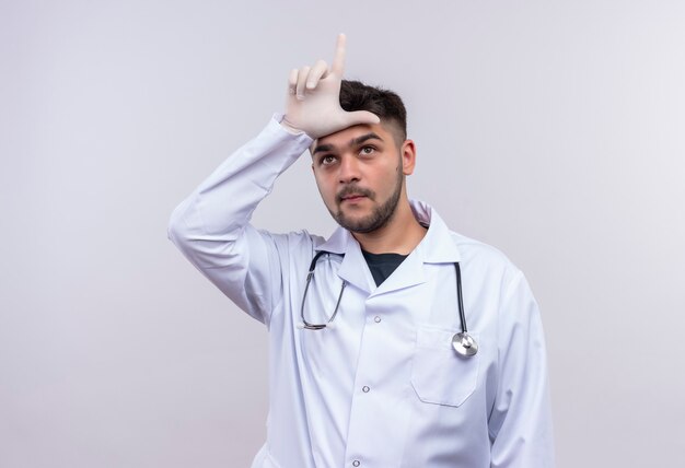 흰색 의료 가운 흰색 의료 장갑과 흰 벽 위에 서있는 느슨한 기호를 보여주는 청진기를 입고 젊은 잘 생긴 의사
