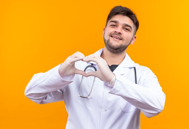 흰색 의료 가운 흰색 의료 장갑과 청진기를 입고 젊은 잘 생긴 의사, 오렌지 벽 위에 서있는 손으로 사랑 기호를 보여주는