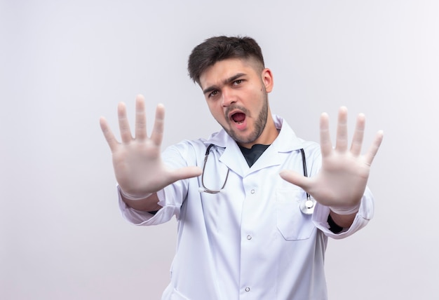 Young handsome doctor wearing white medical gown white medical gloves and stethoscope showing stop sign with hands standing over white wall