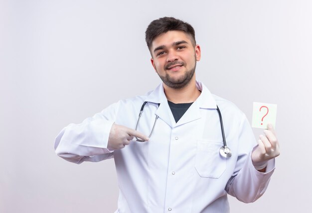 Молодой красивый врач в белом медицинском халате, белые медицинские перчатки и стетоскоп, улыбаясь, указывая на вопросительный знак в руке, стоящей над белой стеной