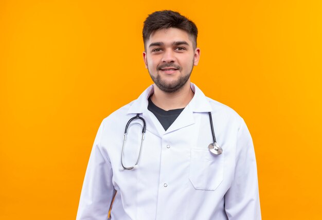 Молодой красивый врач в белом медицинском халате, белые медицинские перчатки и стетоскоп, улыбаясь, стоя над оранжевой стеной