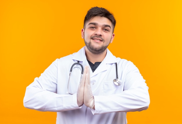 Young handsome doctor wearing white medical gown white medical gloves and stethoscope thankfully greets standing over orange wall