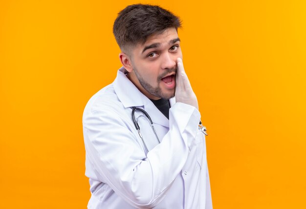 Young handsome doctor wearing white medical gown white medical gloves and stethoscope whispering