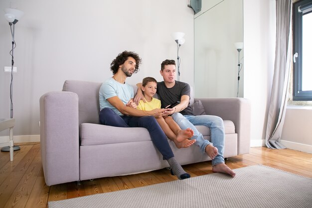 Young handsome gay couple and their son watching TV show at home, sitting on couch in living room, hugging, using remote control, looking away. Family and home entertainment concept