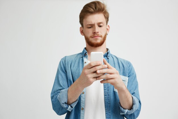 インターネットでサーフィンをして携帯電話の画面を見て眉をひそめている若いハンサムな男。