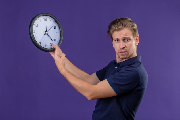 Бесплатное фото Молодой красивый парень держит часы, глядя в камеру с смущенным выражением лица, стоящим на фиолетовом фоне