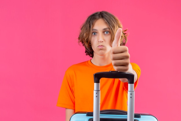 Бесплатное фото Молодой красивый парень в оранжевой футболке держит дорожный чемодан, глядя в камеру, удивлен, показывая большие пальцы вверх стоя